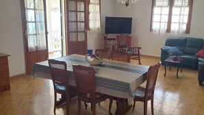 Un coin repas avec une table dressée pour quatre personnes, entourée de chaises en osier. Un téléviseur est fixé au mur, et un canapé est placé dans l’espace salon.