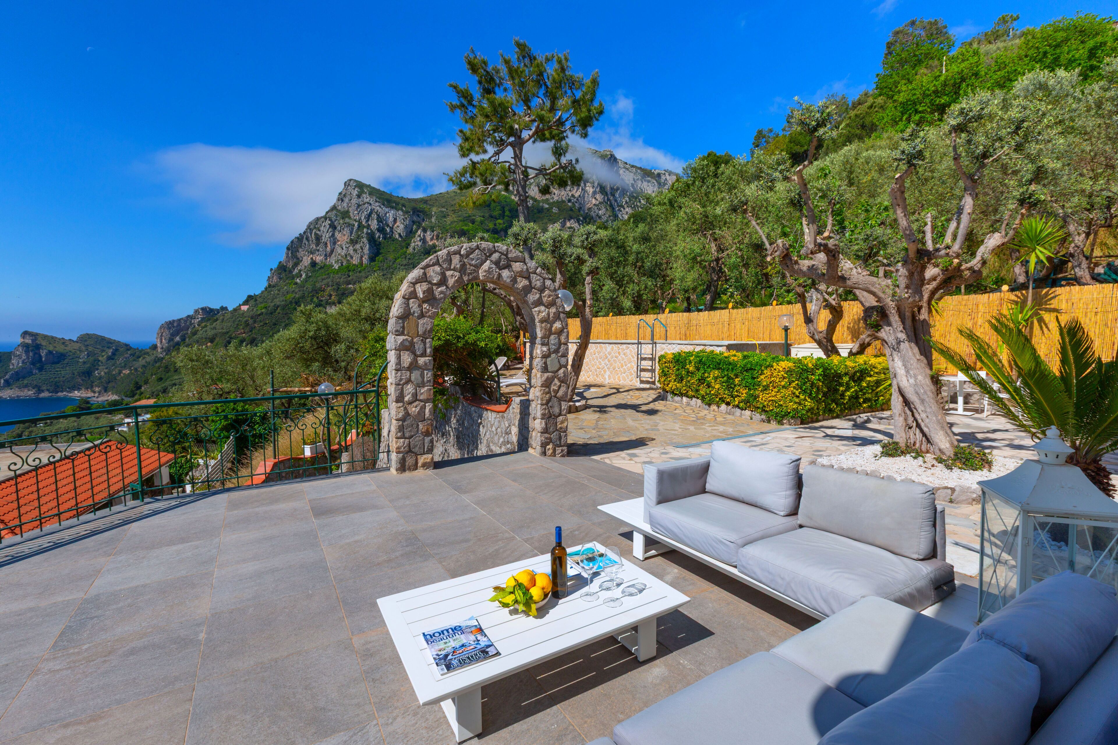 A terrace with a stone archway, outdoor seating, and a view of the sea and mountains.