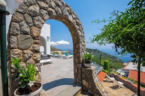 Un arc en pierre mène à une terrasse dotée de sièges extérieurs et offrant une vue sur la mer.