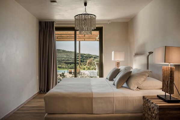 A bedroom with a large bed, a chandelier, a nightstand, and a view of a landscape through a large window.