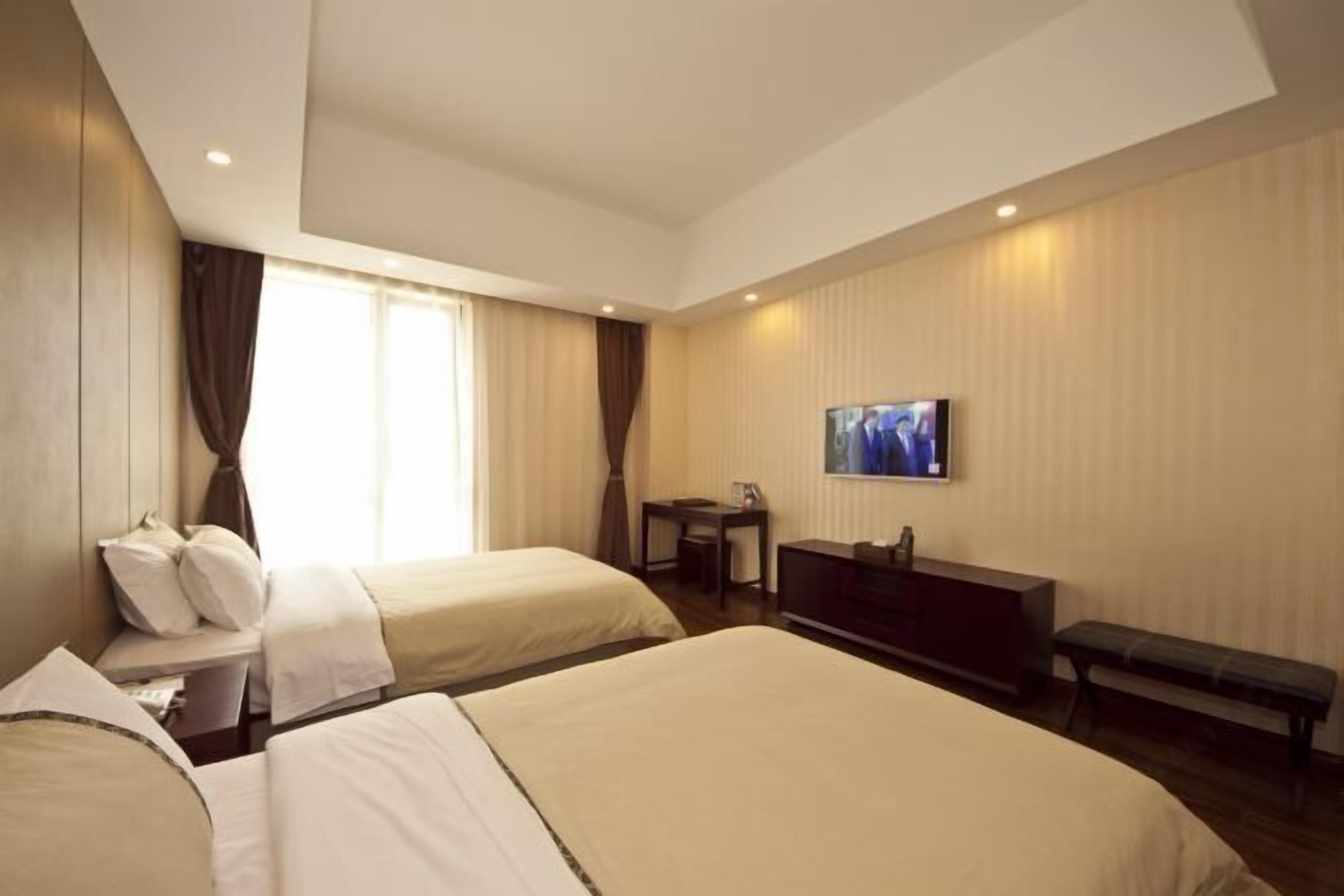 A hotel room with two beds, a desk, a television, and a bench.