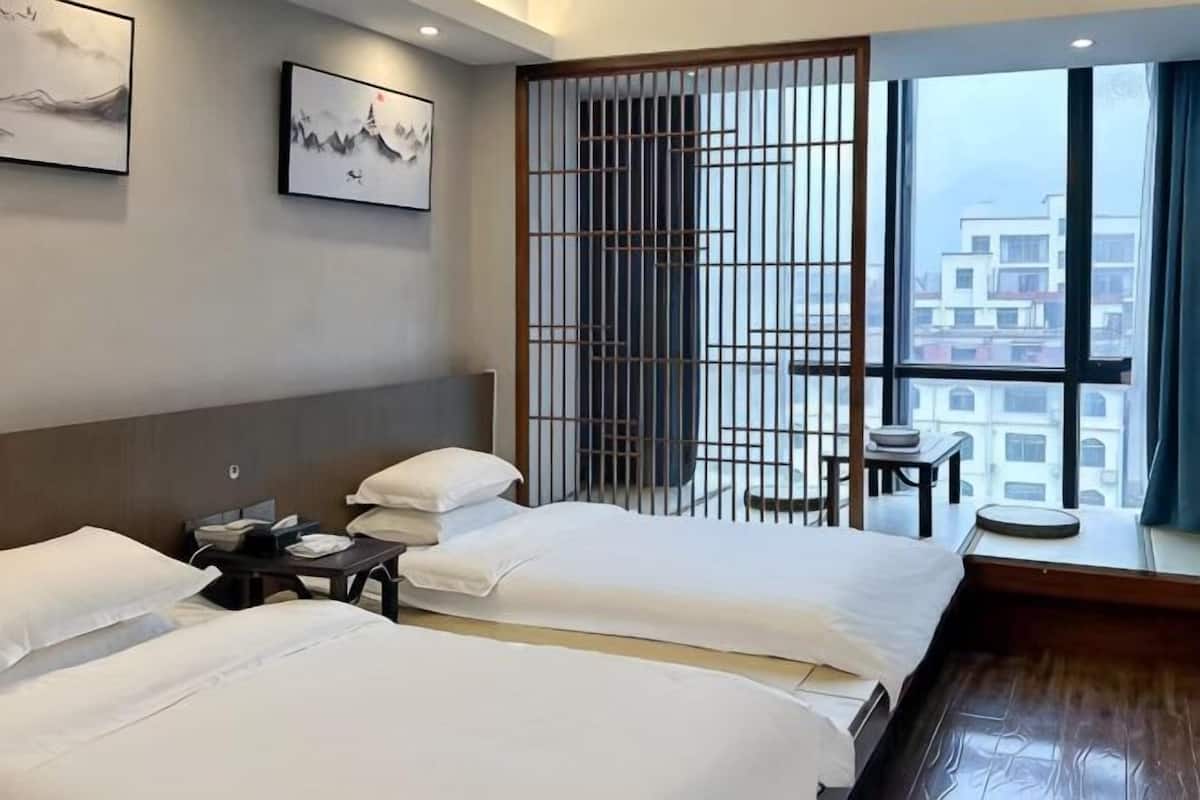 Une chambre d’hôtel avec deux lits, une tête de lit en bois, une fenêtre donnant sur des immeubles et une porte coulissante avec un moustiquaire métallique.