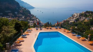 A swimming pool with lounge chairs and umbrellas, surrounded by buildings and a view of the sea.