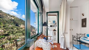 A balcony with a view of a hillside village, a dining area with a table and chairs, and a kitchen visible through an open door.