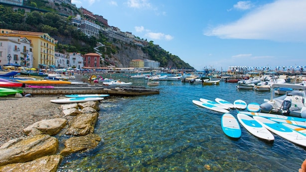 A coastal town with boats docked along the shore, buildings on a hillside, and clear blue water.
