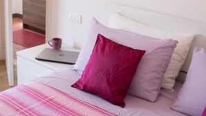 Una cama bien hecha con una computadora portátil sobre una mesita de noche blanca, un almohán morado y una manta a rayas rojas y blancas.