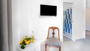 Una habitación con una televisión de pantalla plana montada en la pared, una silla de madera con un cojín a rayas, una pequeña mesa blanca con una botella y limones, y una puerta que da a un pasillo con un estampado azul y blanco a cuadros.