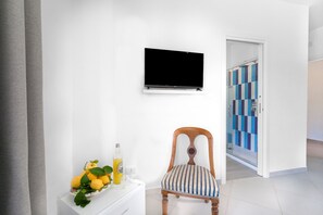 Una habitación con una televisión de pantalla plana montada en la pared, una silla de madera con un cojín a rayas, una pequeña mesa blanca con una botella y limones, y una puerta que da a un pasillo con un estampado azul y blanco a cuadros.