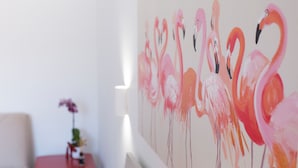 Une pièce avec un mur blanc et un tableau représentant des flamants roses. On y trouve une petite table avec un vase de fleurs et une chaise.