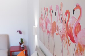 Een kamer met een witte muur en een schilderij van flamingo's. Er staat een kleine tafel met een bloevas en een stoel.