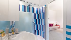 Une salle de bain moderne avec une grande baignoire blanche, une douche en verre avec des carreaux bleus et blancs, et un canapé blanc dans le salon attenant.