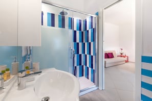 Een moderne badkamer met een groot, wit bad, een douche met glazen wanden en blauw-witte tegels, en een witte bank in de aangrenzende woonruimte.