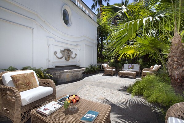A patio with wicker furniture, a stone fountain, and lush greenery.