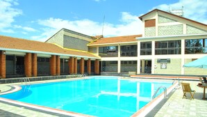 Ein Schwimmbecken mit Fliesen umrandet, einem überdachten Sitzbereich und einem Gebäude mit zahlreichen Fenstern und einer Ziegel Fassade.