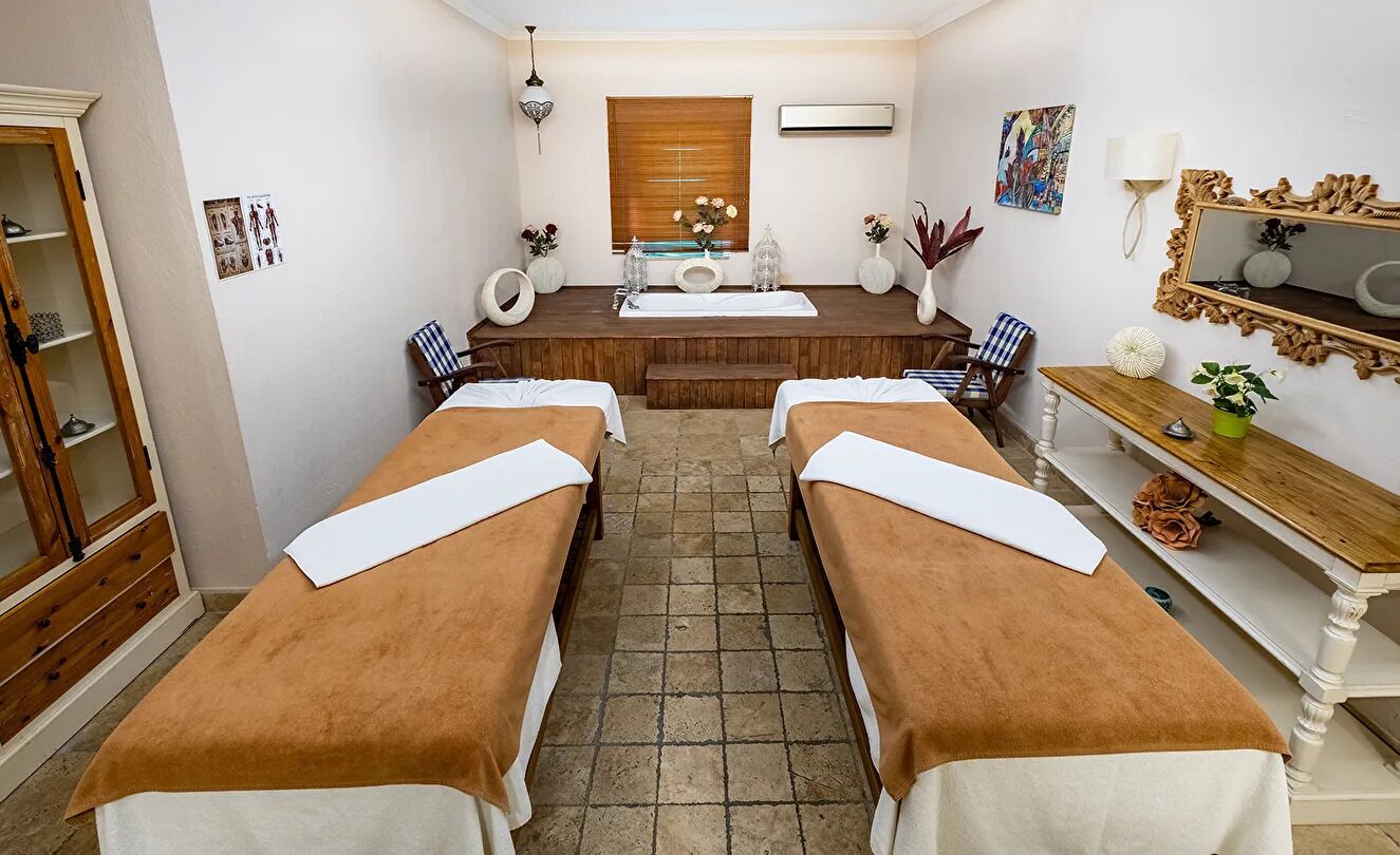 A spa room with two massage tables, a wooden bench, a mirror, and a window with blinds.