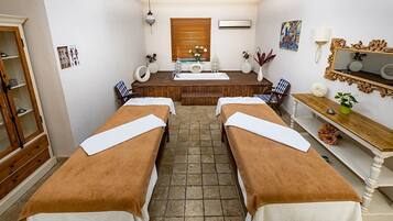 A spa room with two massage tables, a wooden bench, a mirror, and a window with blinds.