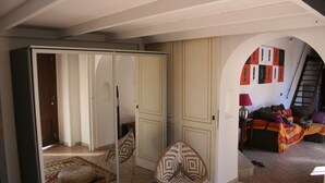 Ein Zimmer mit Schiebetürenschrank, einem Schminktisch mit Spiegel, einem gemusterten Sessel und einer Couch mit farbenfroher Decke.