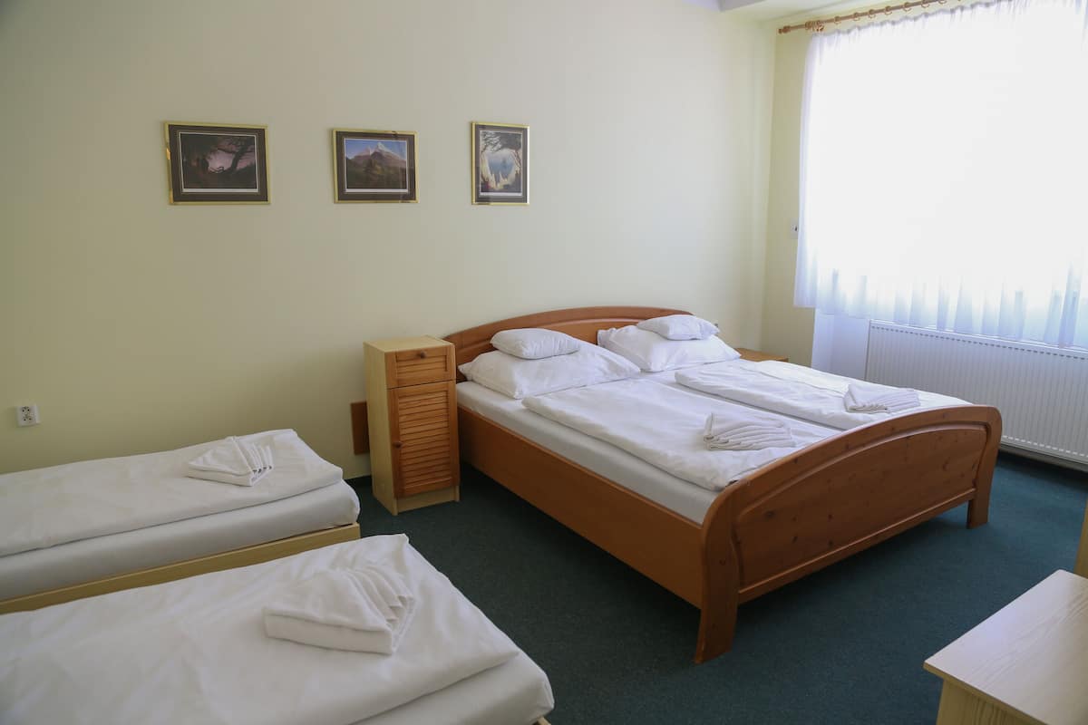 Una habitación con dos camas individuales y una cama doble, todas ordenadas con ropa de cama blanca. Hay tres cuadros en la pared y una ventana con cortinas traslúcidas.