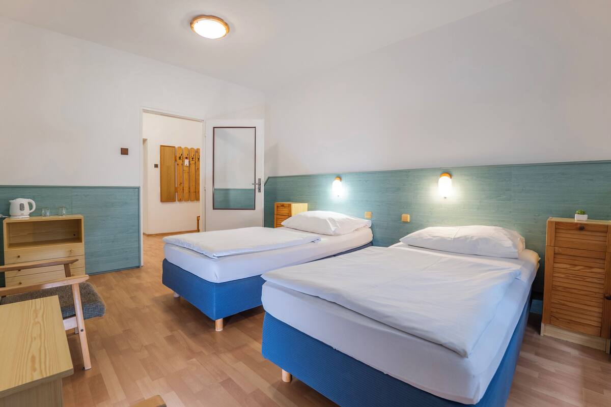 Habitación de hotel con dos camas individuales, un escritorio de madera, una silla, una mesita con una tetera y un armario.