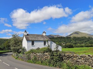 A white house with a stone wall and a stone fence, surrounded by greenery and mountains in the background. - Ellis Howe (Coniston)