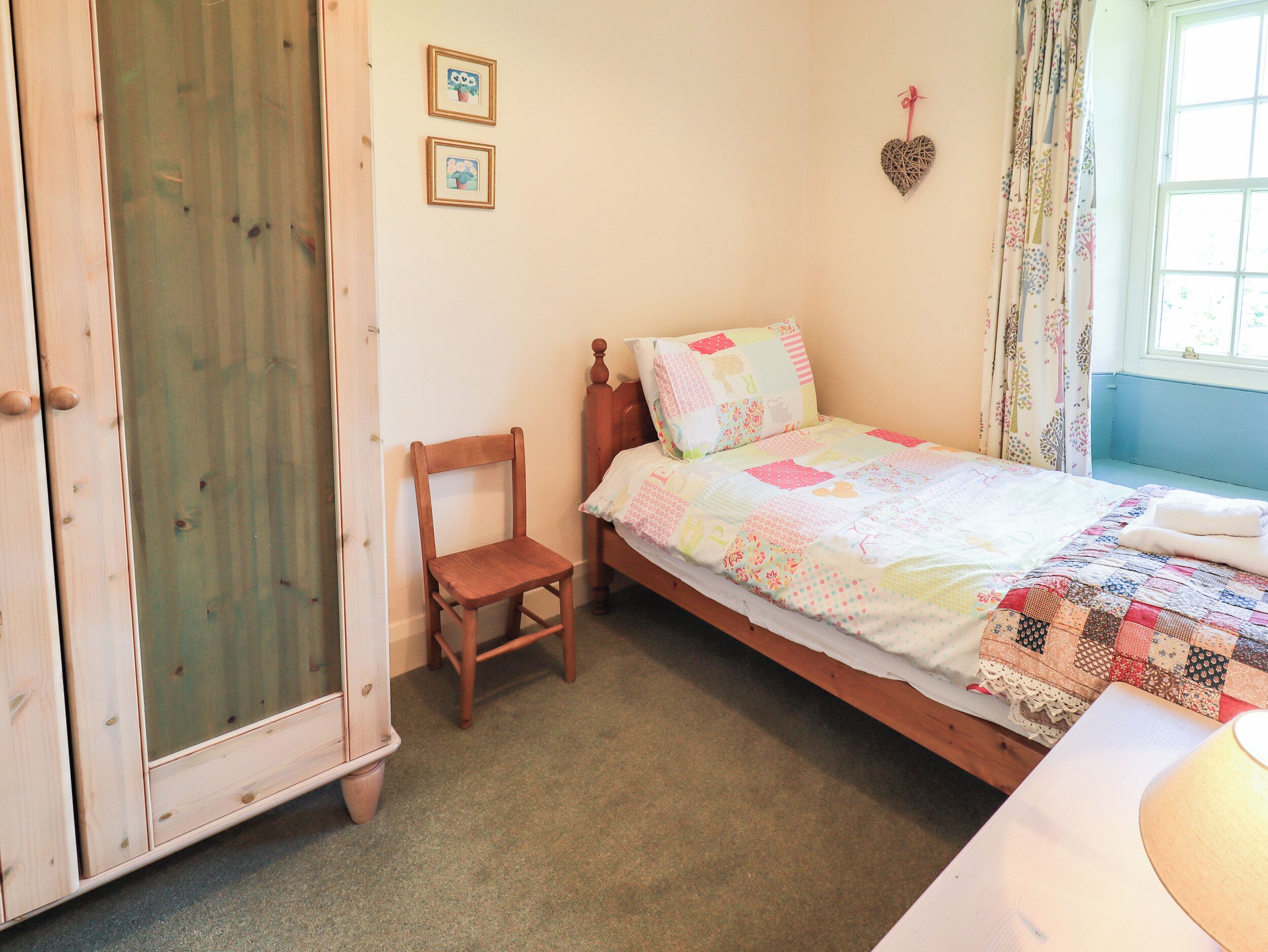Un dormitorio con cama de madera, una pequeña silla de madera, una ventana con cortinas y dos cuadros en la pared.