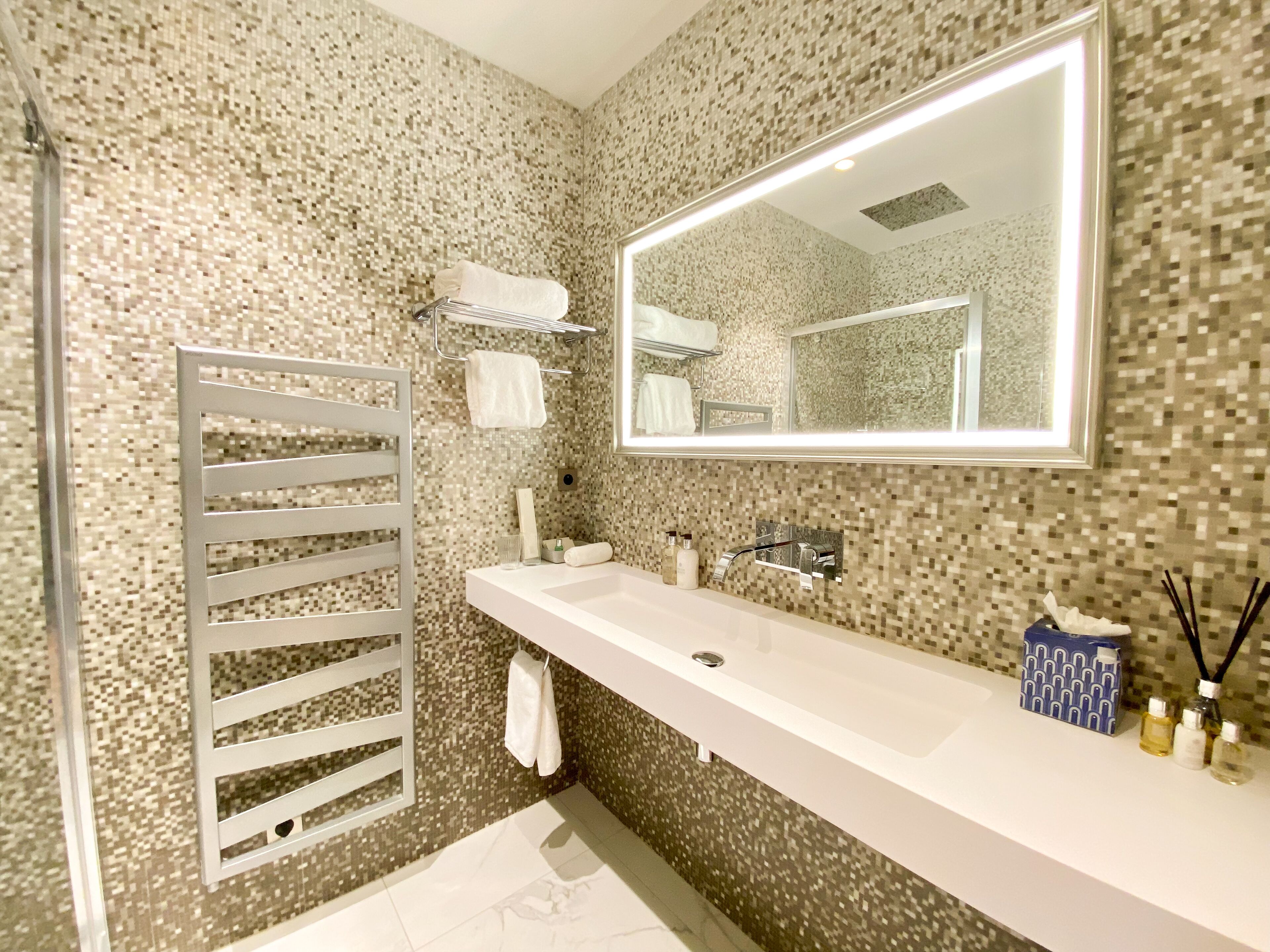 A modern bathroom with a tiled mosaic wall, a large mirror, a white sink, and a towel rack.
