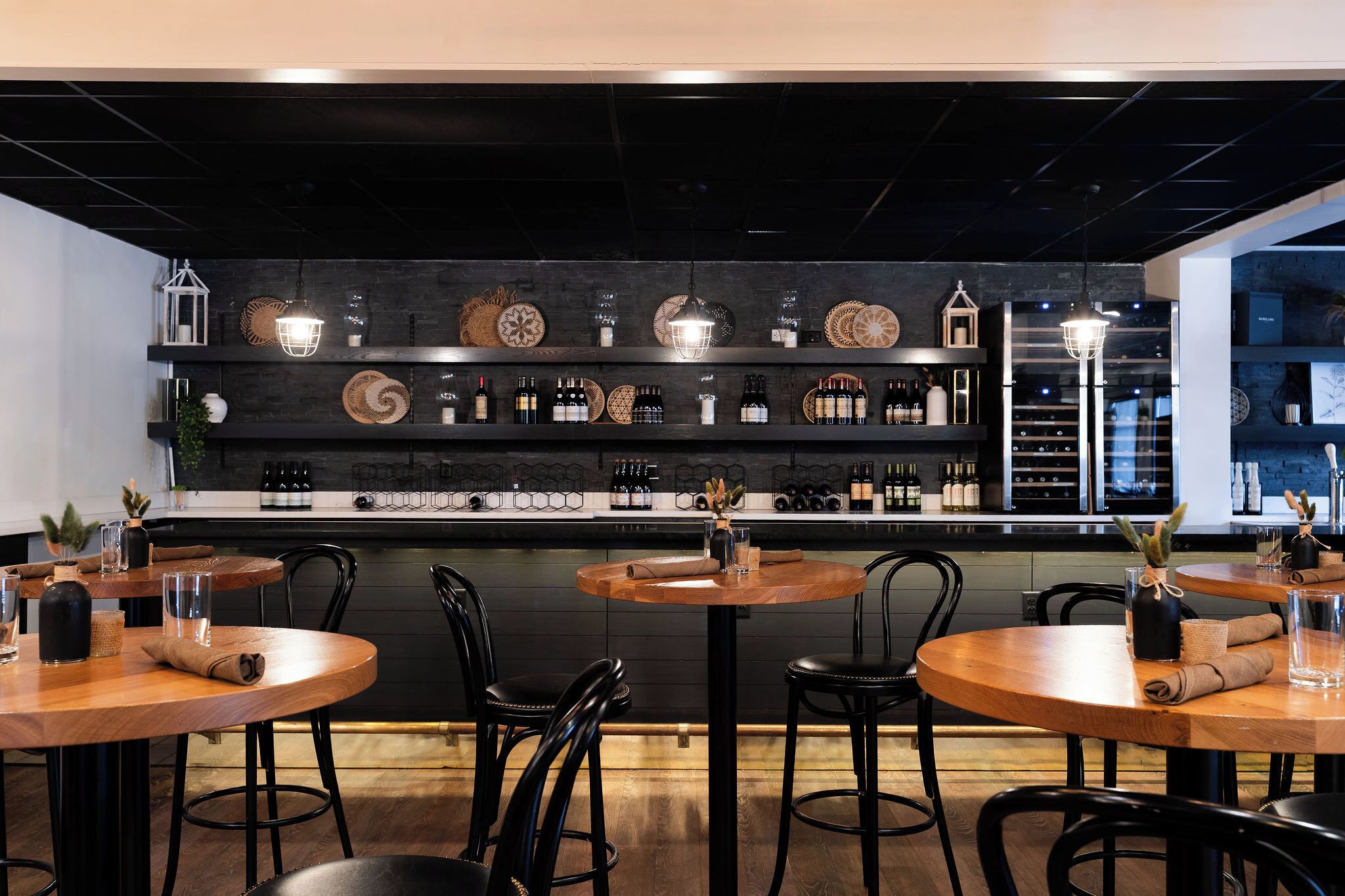 Een moderne bar met ronde houten tafels, zwarte stoelen en diverse flessen en decoratieve objecten op planken.