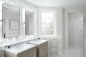A modern bathroom with two sinks, large mirrors, a window, and a shower with a marble herringbone pattern.