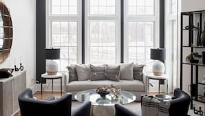 A modern living room with a large window, a chandelier, a sofa, two armchairs, a coffee table, and a side table with a lamp. - The Federal (Brunswick)
