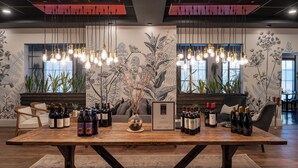 A wooden table with wine bottles and a framed certificate, surrounded by chairs and hanging light fixtures in a modern interior with large windows and plant decorations. - The Federal (Brunswick)
