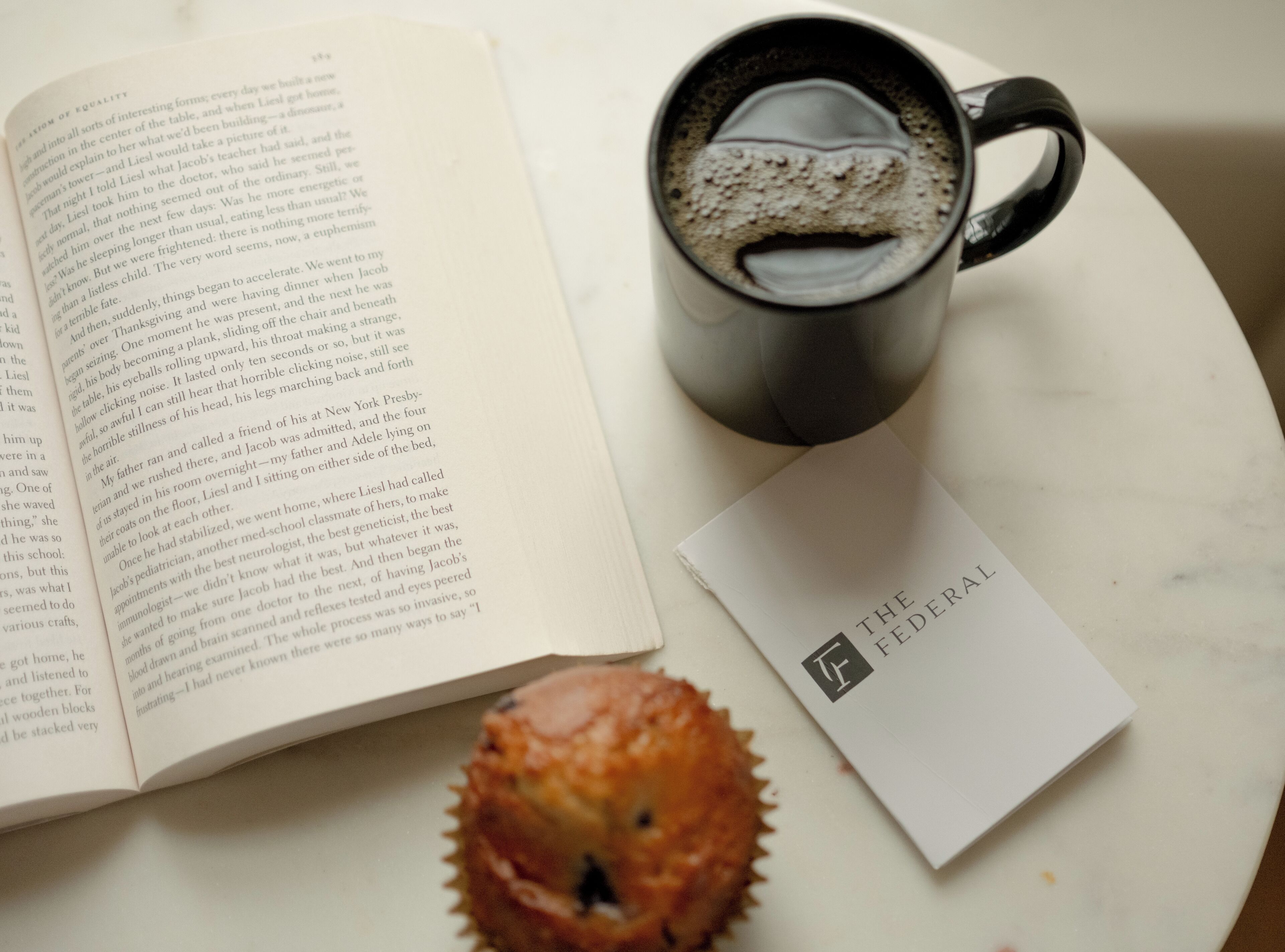 Un libro abierto, una taza de café y un muffin sobre una superficie de mármol.