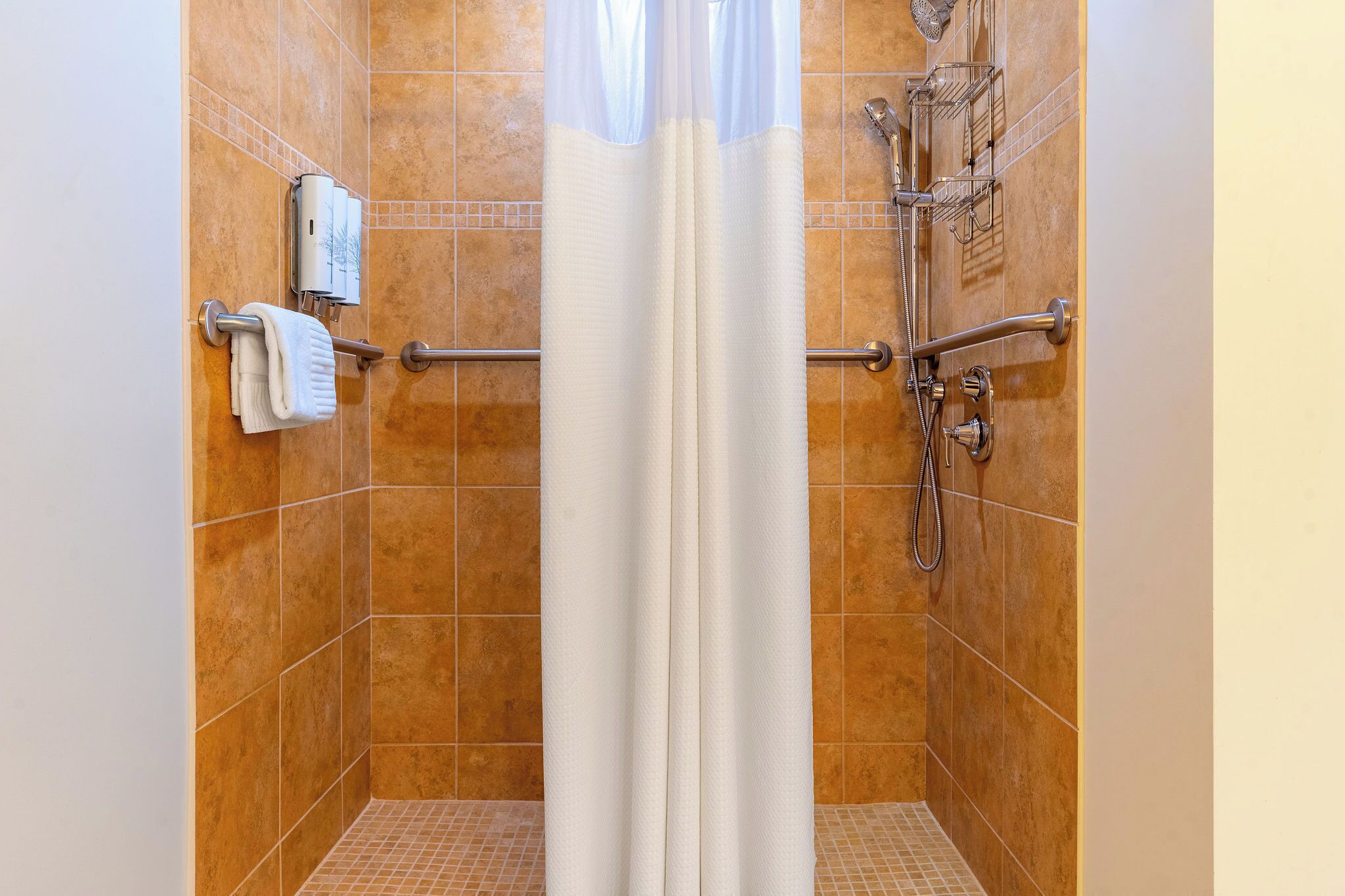 A shower stall with a white curtain, a handheld showerhead, a wall-mounted towel dispenser, and a handheld shower head.