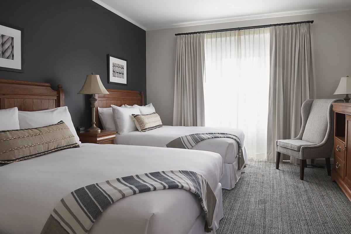 A hotel room with two beds, a wooden headboard, a lamp, a chair, and a large window with curtains.