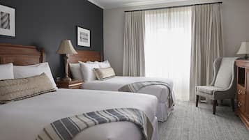 A hotel room with two beds, a wooden headboard, a lamp, a chair, and a large window with curtains.