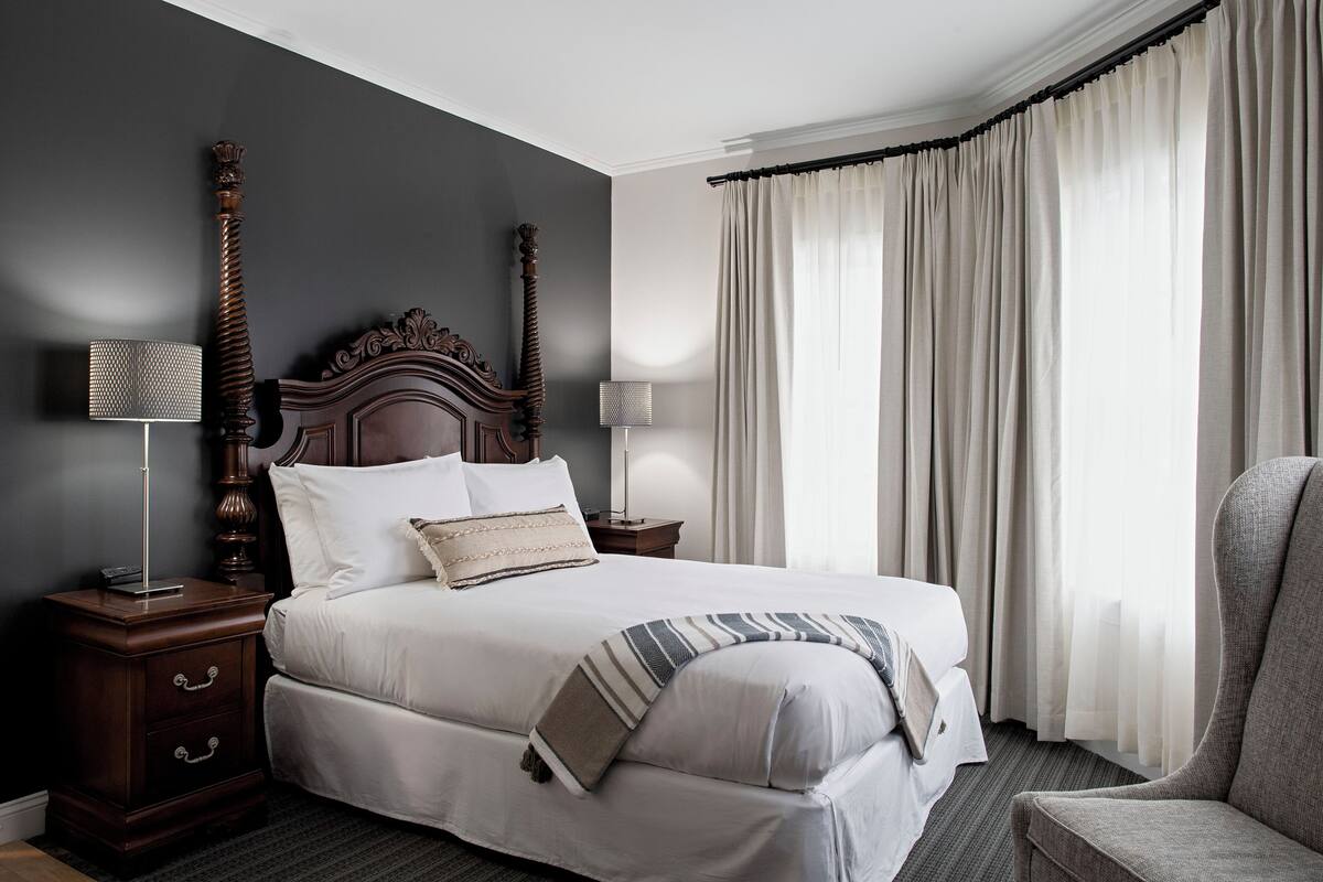 A neatly made bed with white linens, a dark wooden headboard, and a striped throw blanket. There are two bedside tables with lamps, and a chair is positioned near the window.
