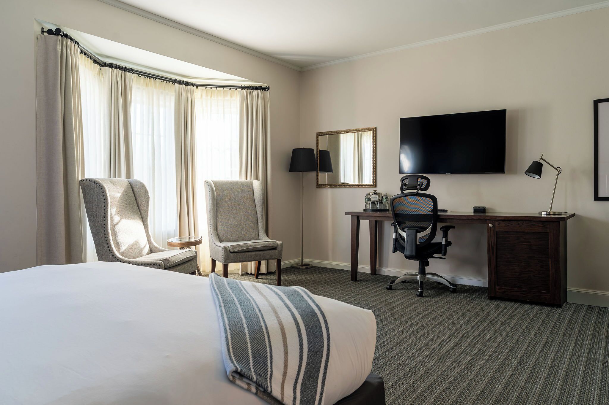 A hotel room with a large bed, two armchairs, a desk with a chair, a flat-screen TV, and a large mirror.