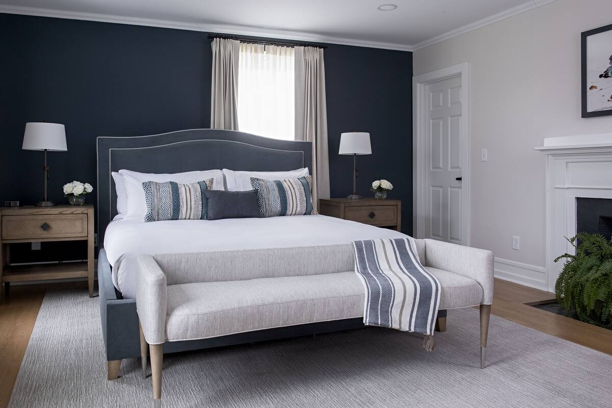 A bedroom with a large bed, two bedside tables, a nightstand, a fireplace, and a window with curtains.