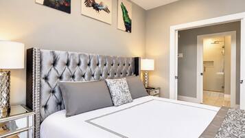 A bedroom with a tufted headboard, a large bed with grey bedding, two bedside tables with lamps, and three wall-mounted artworks.