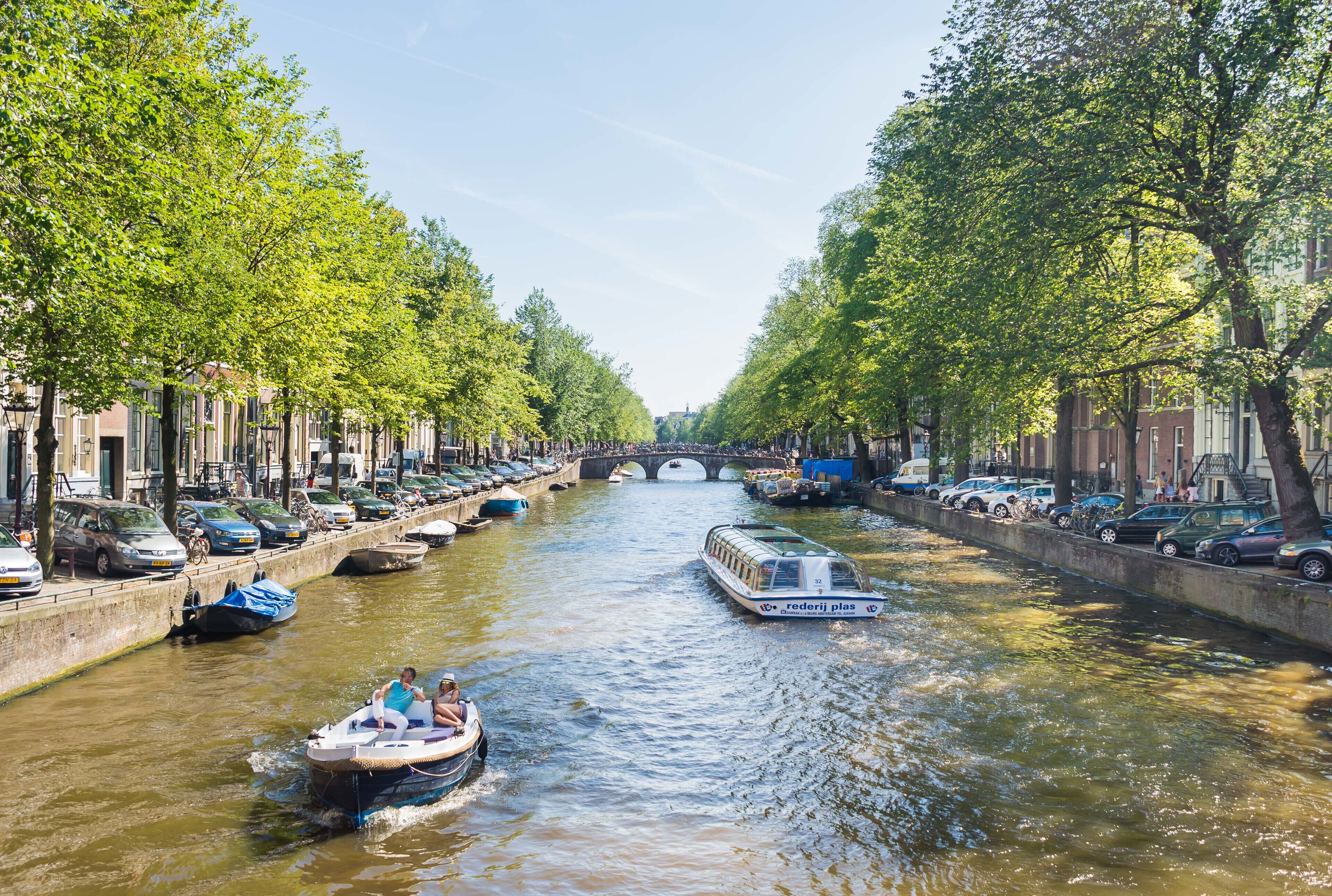 Een gracht met boten, omzoomd door bomen en parkeerauto's aan beide zijden.