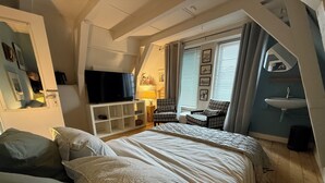 A bedroom with a large bed, a TV on a white stand, a chair, a lamp, and a window with blinds. - BB 1971 Hazenstraat (shared bathroom) (Amsterdam)