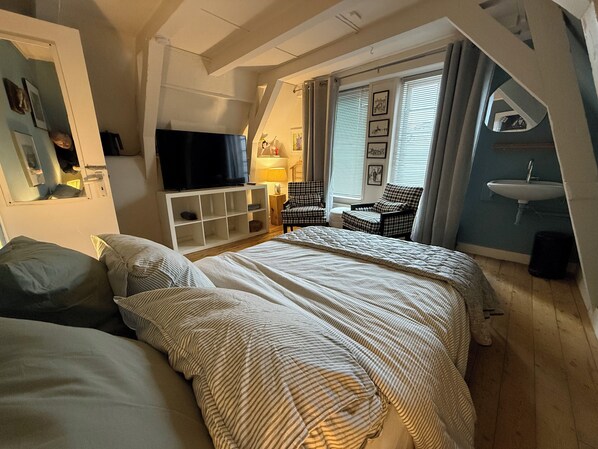 A bedroom with a large bed, a TV on a white stand, a chair, a lamp, and a window with blinds.