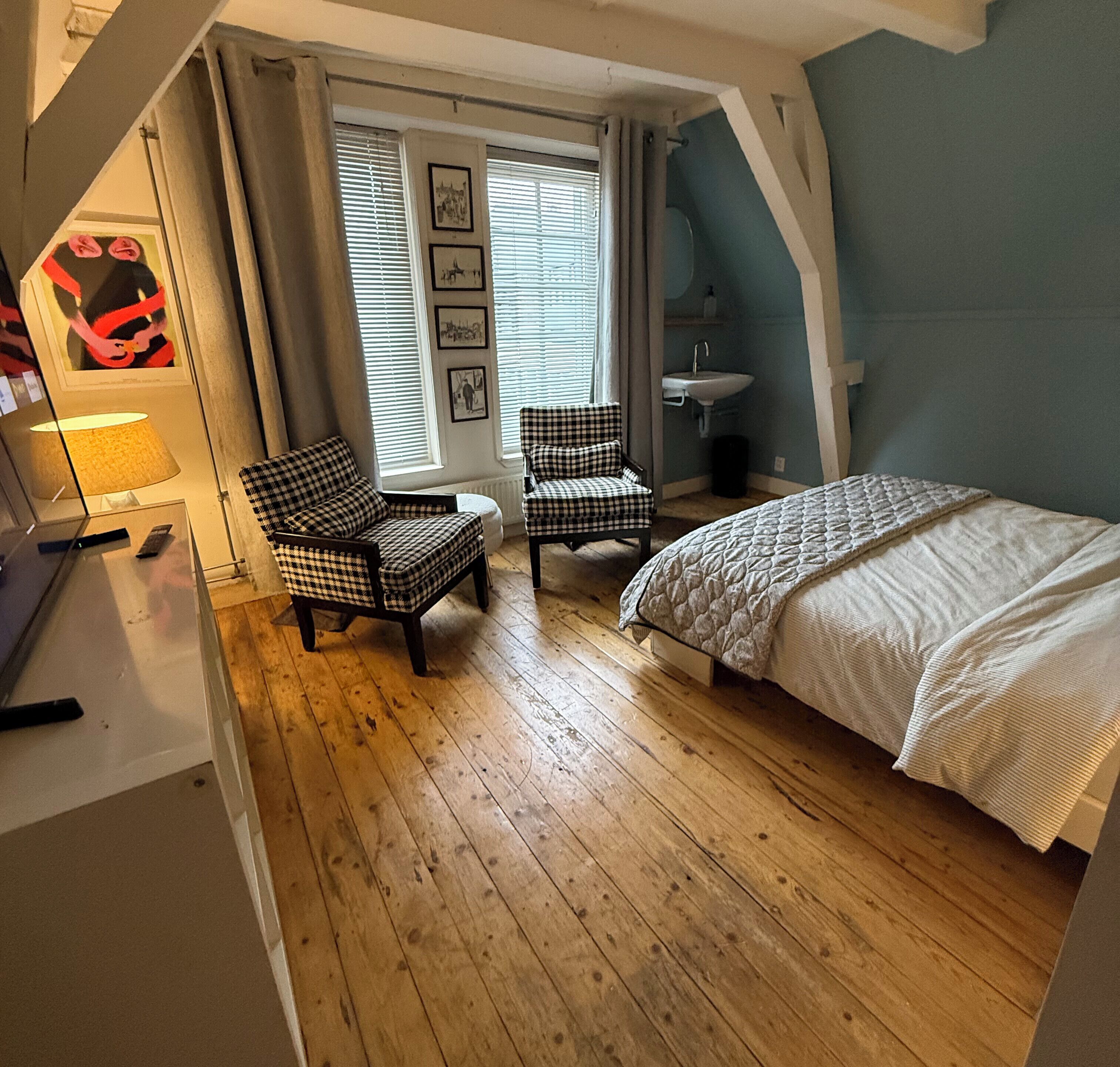 Une chambre à coucher avec un lit, deux chaises à carreaux, une fenêtre avec des stores, une lampe et un lavabo.