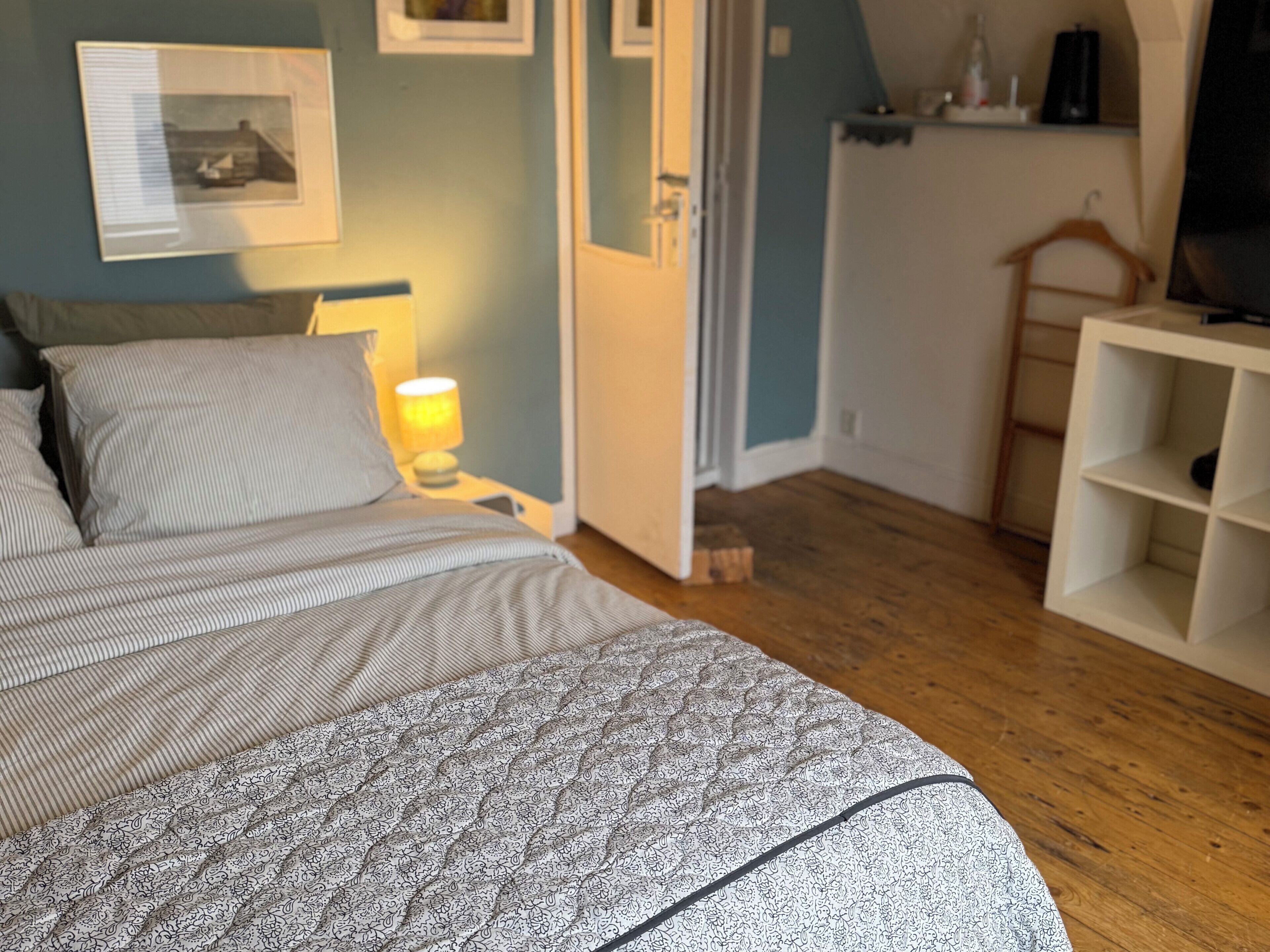 A bedroom with a bed, a nightstand with a lamp, a wooden floor, a door, and a white shelving unit.