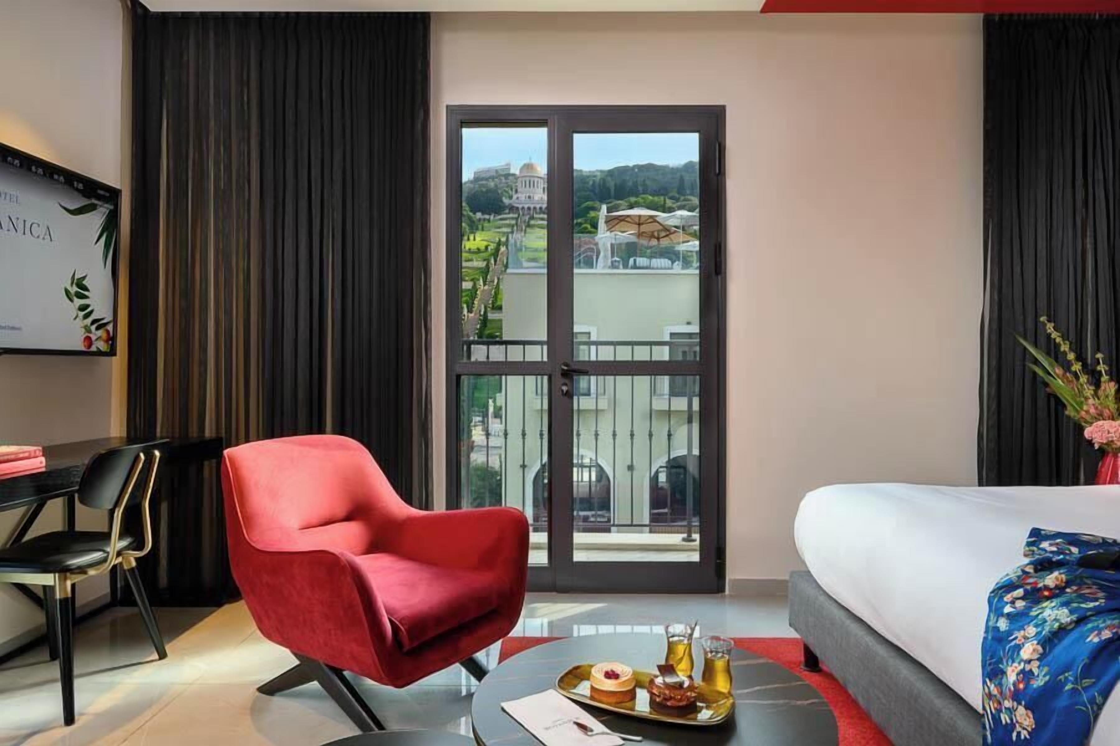 Ein Hotelzimmer mit einem Bett, einem roten Sessel, einem Esstisch mit einer Platte mit Speisen und Blick auf ein Gebäude durch eine Glastür.