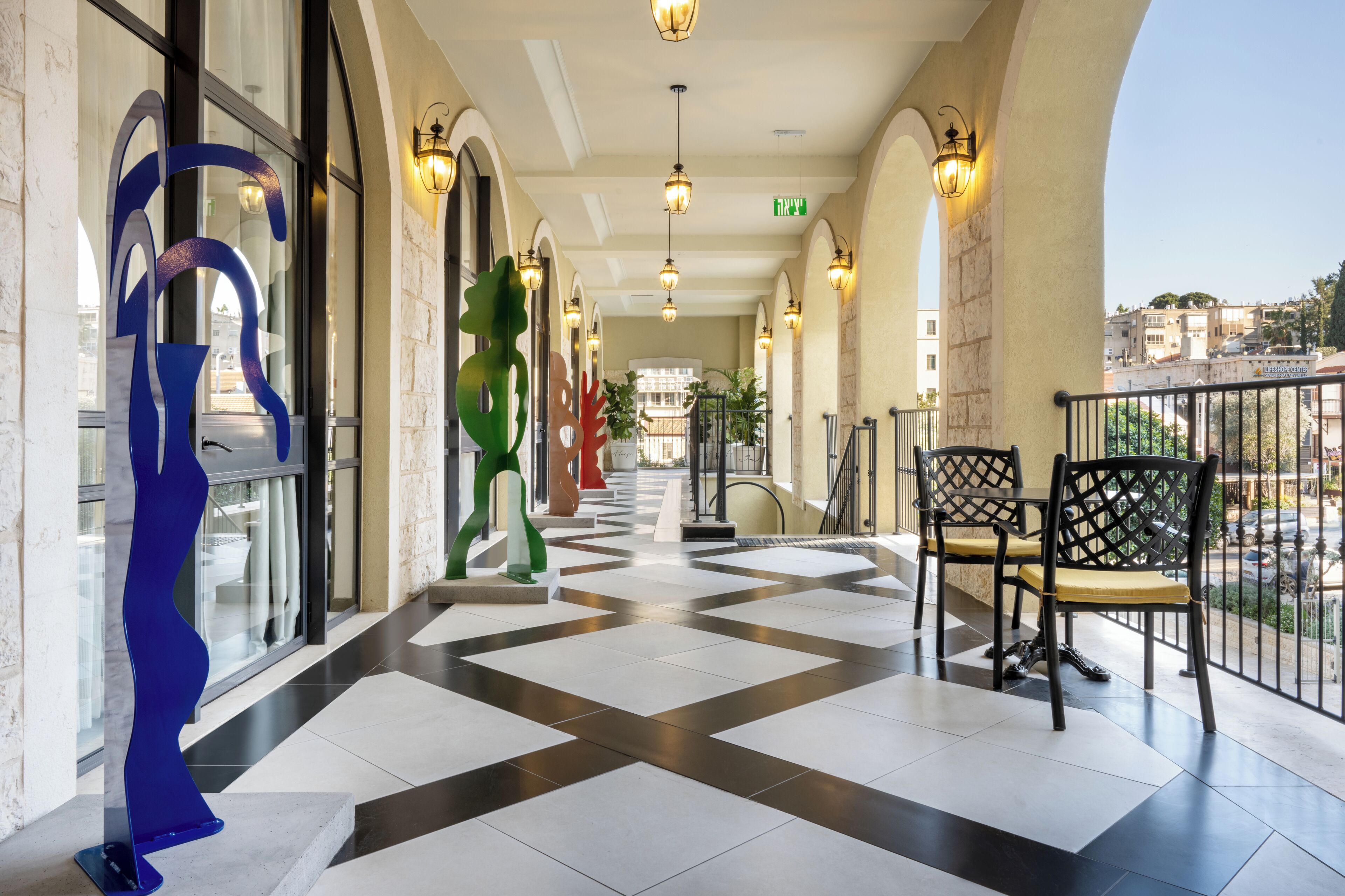 A hotel corridor with a checkered floor, decorative sculptures, and a balcony with chairs.