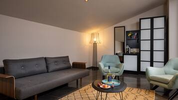 A modern living room with a grey sofa, two light blue armchairs, a small round coffee table with snacks, a floor lamp, and a bookshelf with decorative items.
