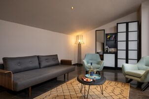 Una sala moderna con un sofá gris, dos sillones azul claro, una mesita redonda con aperitivos, una lámpara de pie y una estantería con objetos decorativos.