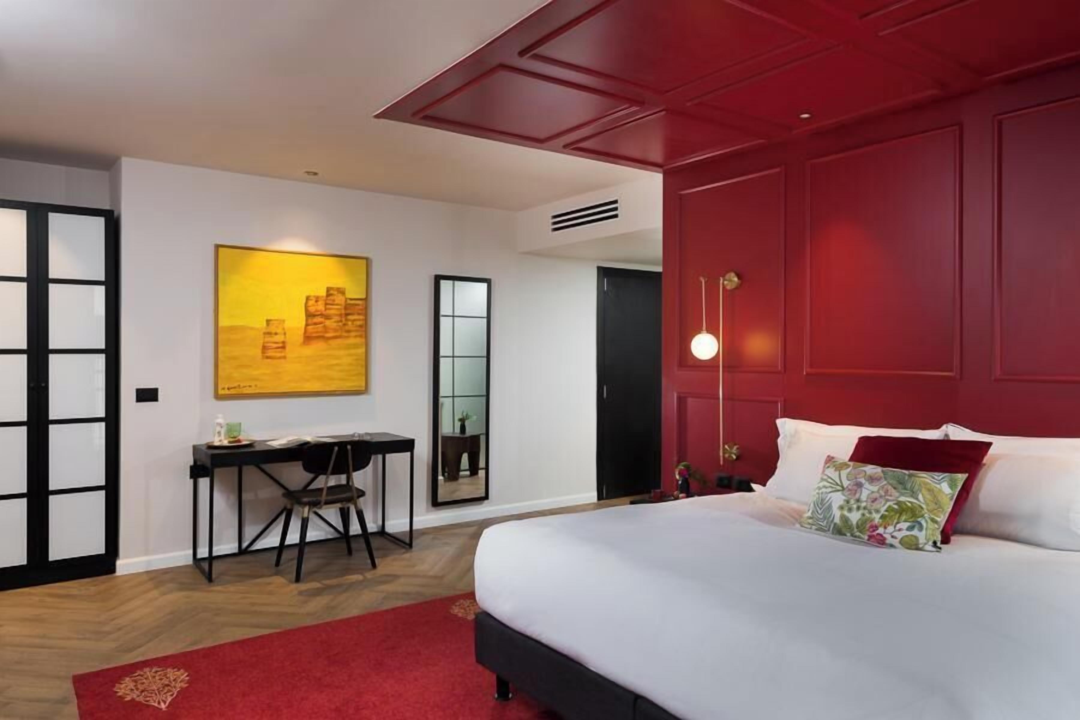 A hotel room with a large bed, a desk with a chair, a red accent wall, and a painting on the wall.