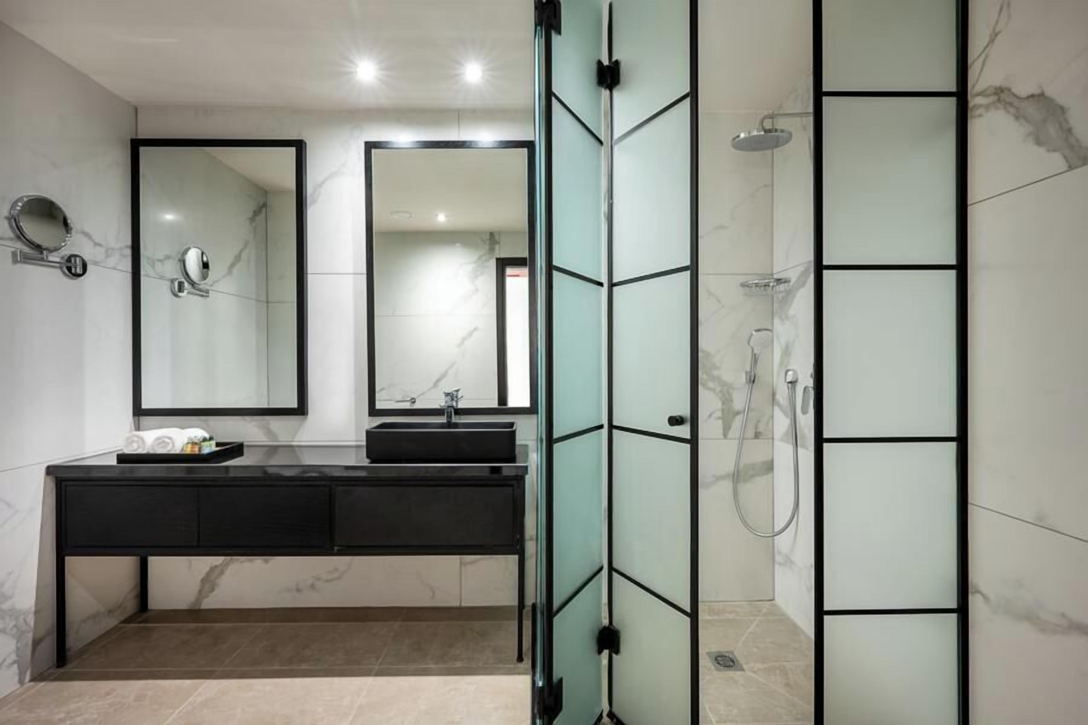 Un baño moderno con tocador negro, dos espejos, cabina de ducha de vidrio y paredes de mármol.