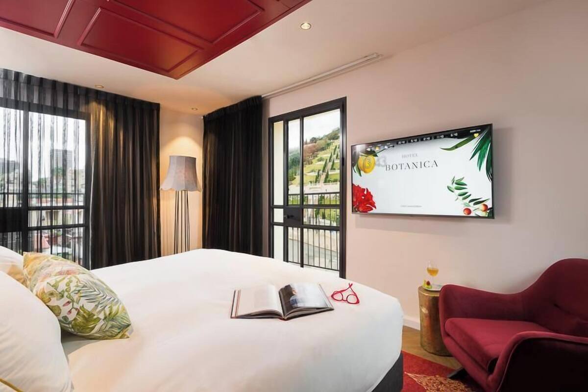 Habitación de hotel con una cama grande, un sillón rojo, un televisor en la pared y una ventana con cortinas.